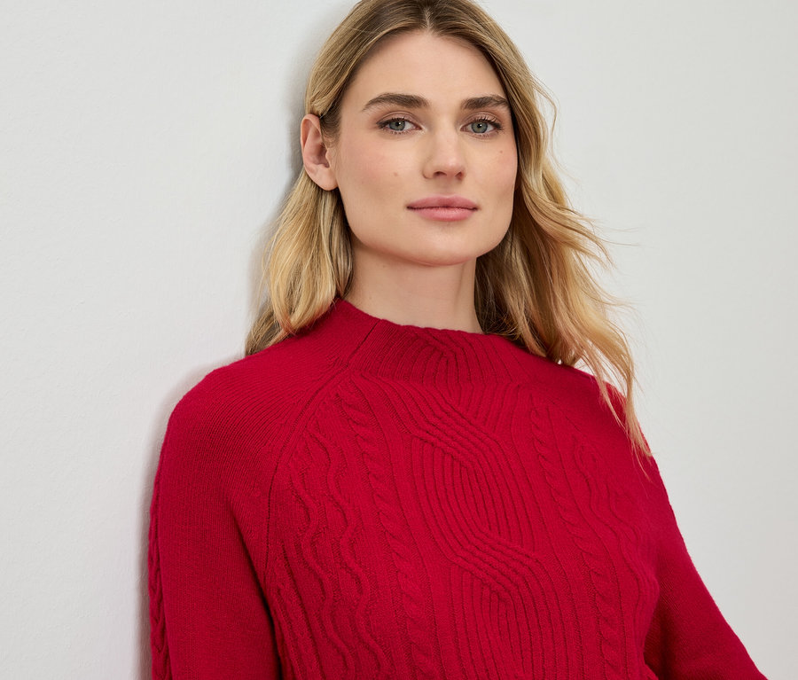 Une femme blonde est appuyée contre un mur et porte un pull rouge.