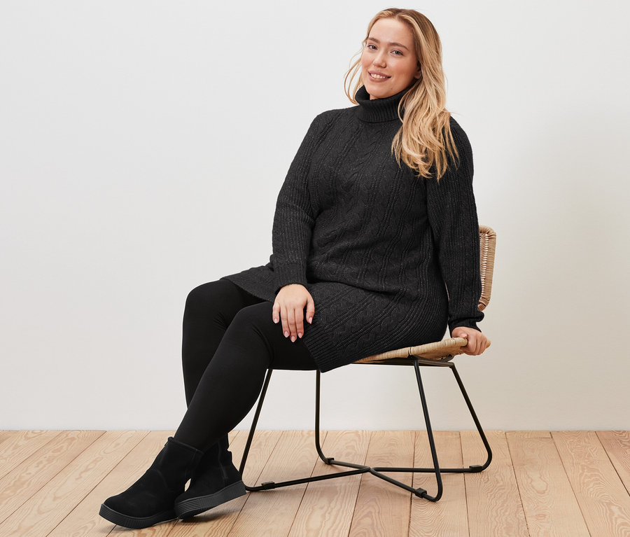 Une femme est assise sur une chaise et porte un long pull en maille noire avec torsades.