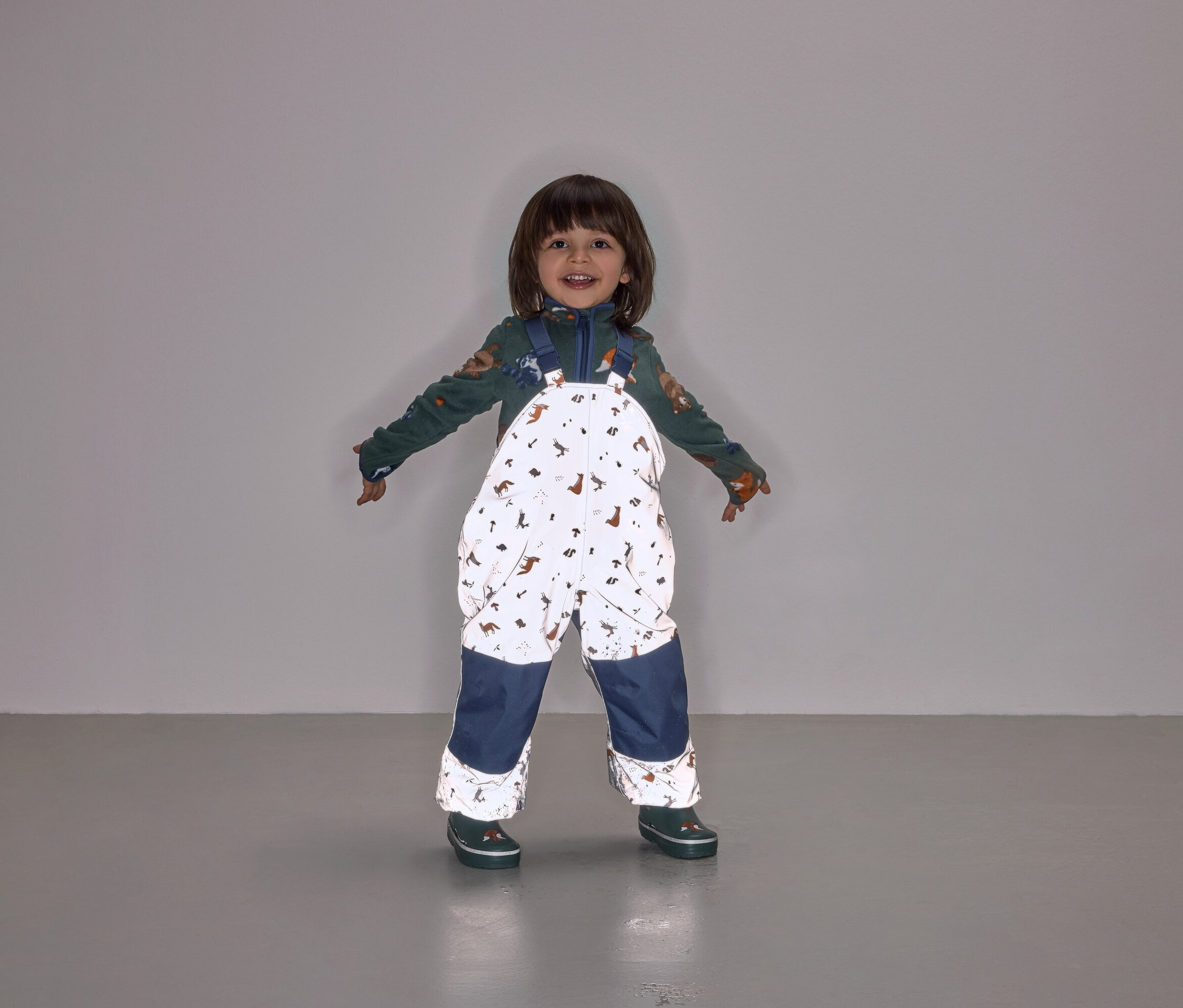 Un enfant souriant se tient debout, les bras écartés, portant une salopette à motifs animaliers avec des jambes bleu marine.