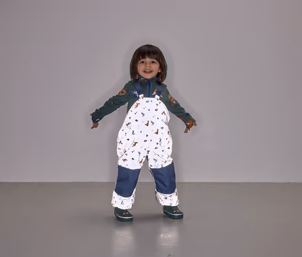 Un enfant souriant se tient debout, les bras écartés, portant une salopette à motifs animaliers avec des jambes bleu marine.