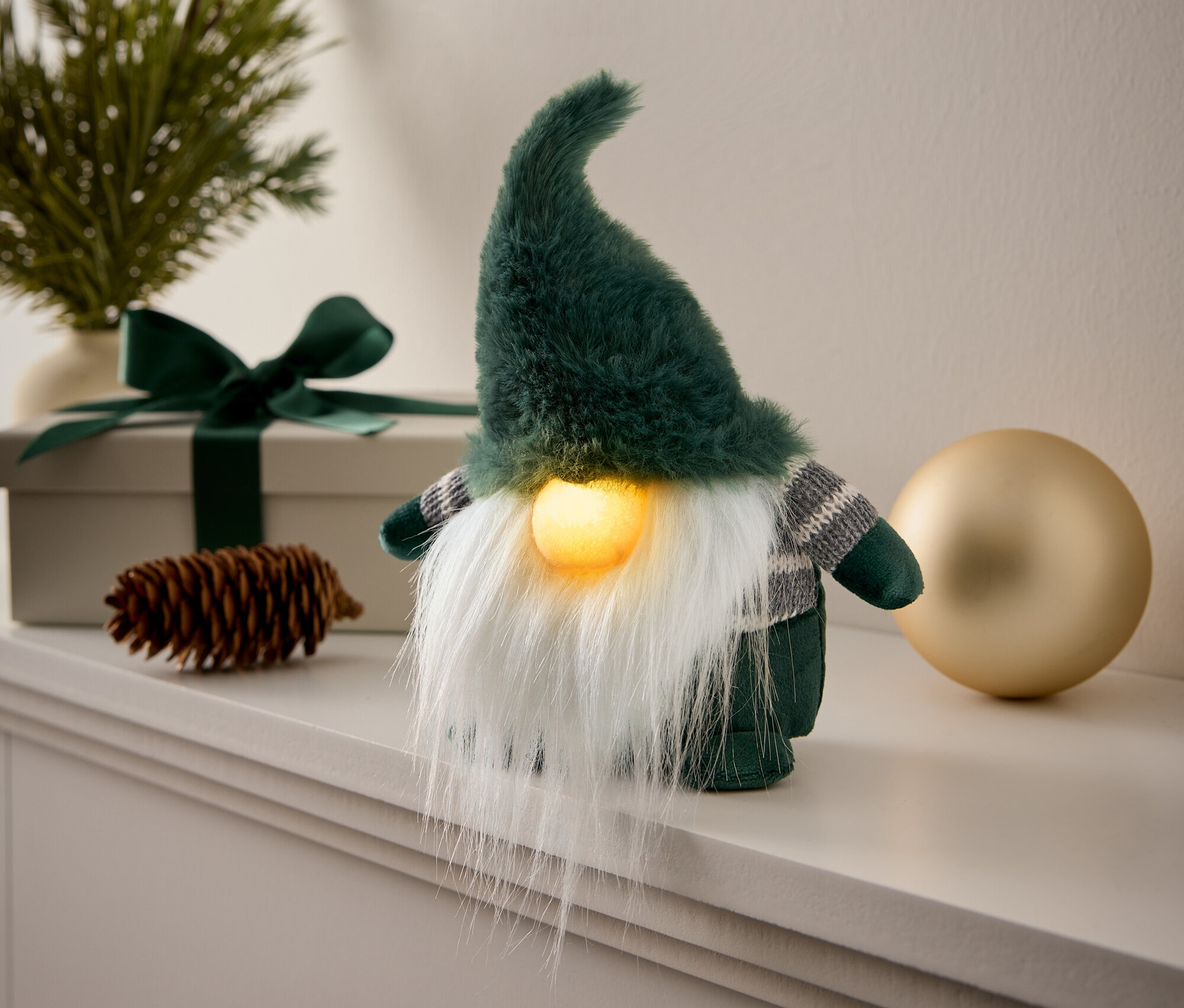 Un gnome décoratif vert avec une barbe blanche est posé sur une cheminée à côté d'une boule dorée.