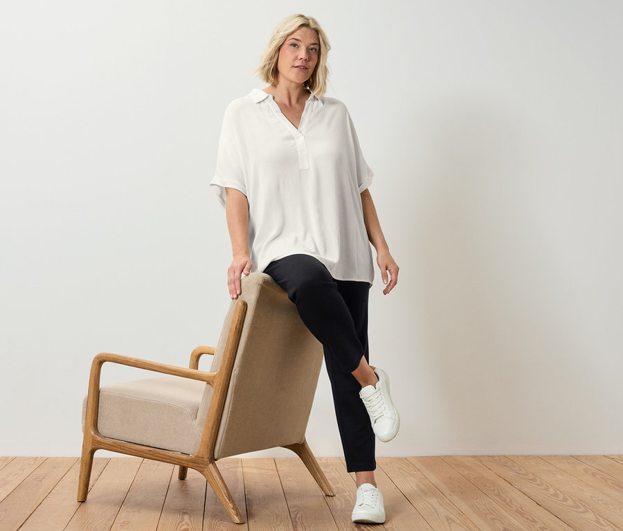 Une femme blonde posant à côté d'un fauteuil beige, portant une blouse crème.