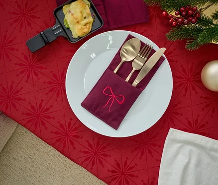 Assiette avec un porte-serviette brodé, des couverts dorés, une poêle à raclette et des décorations de Noël sur une nappe rouge.