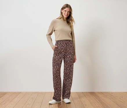 Une femme se tient debout, les mains dans les poches d'un pantalon marron à fleurs, portant un pull beige.