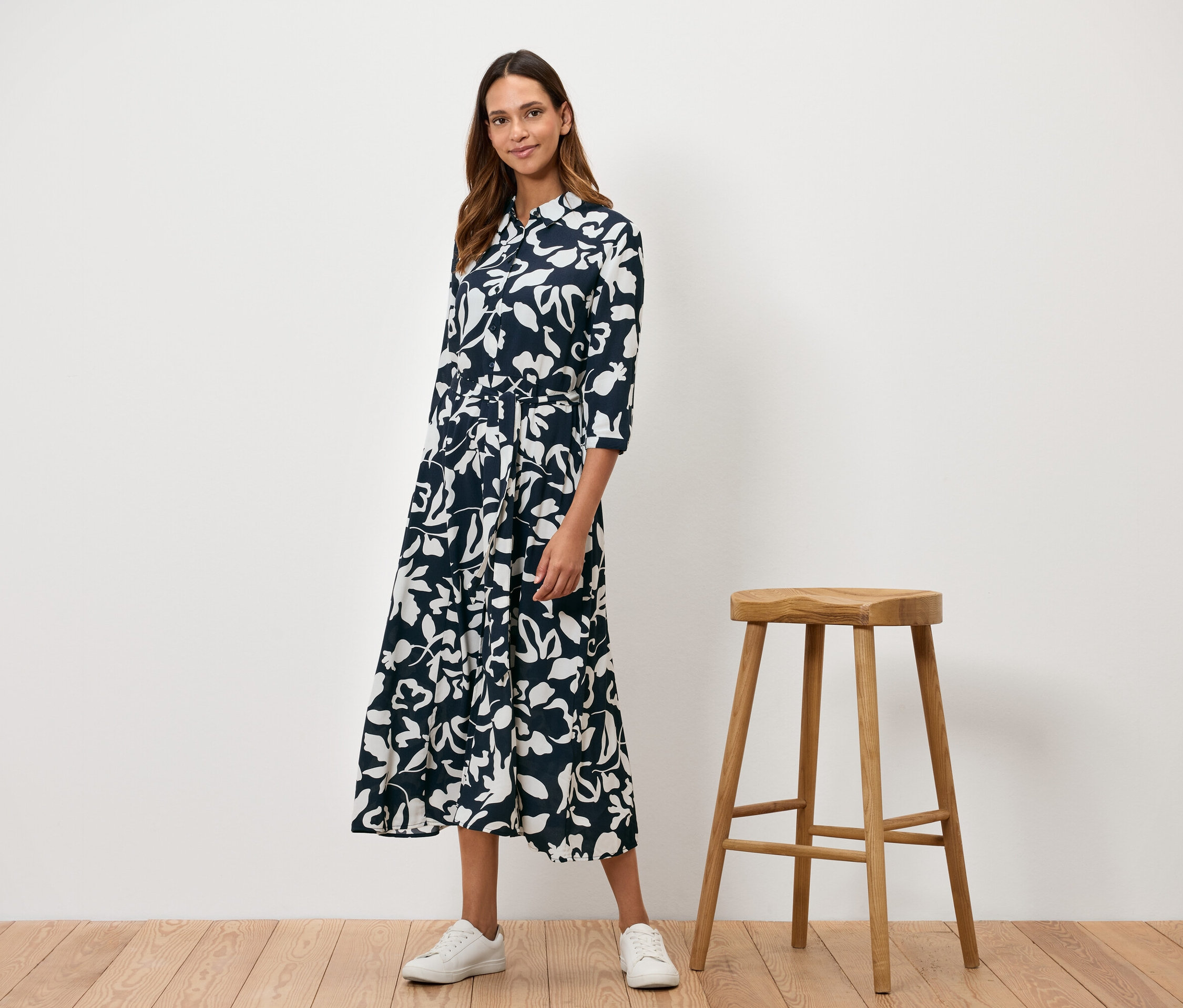 Une femme se tient à côté d'un tabouret en bois et porte une robe bleu marine à fleurs blanches.