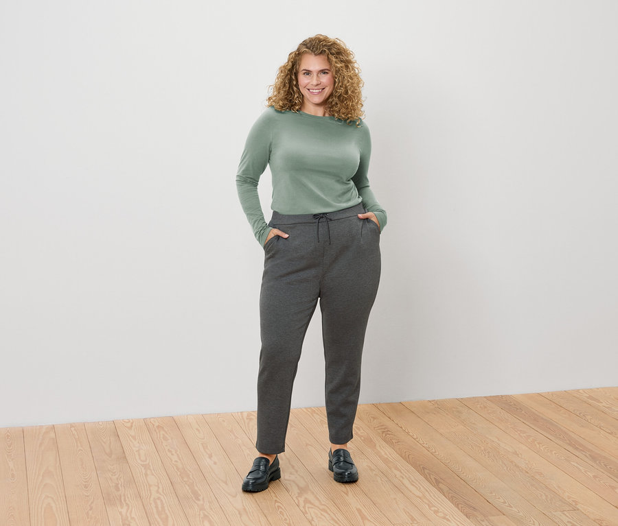 Femme aux cheveux blonds bouclés portant un T-shirt vert à manches longues et un pantalon gris avec ses mains dans les poches.