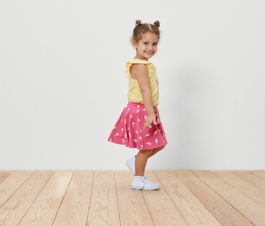 Petite fille souriante posant de côté dans une jupe en jersey pour enfants avec pantalon à motif de glace et un t-shirt jaune.