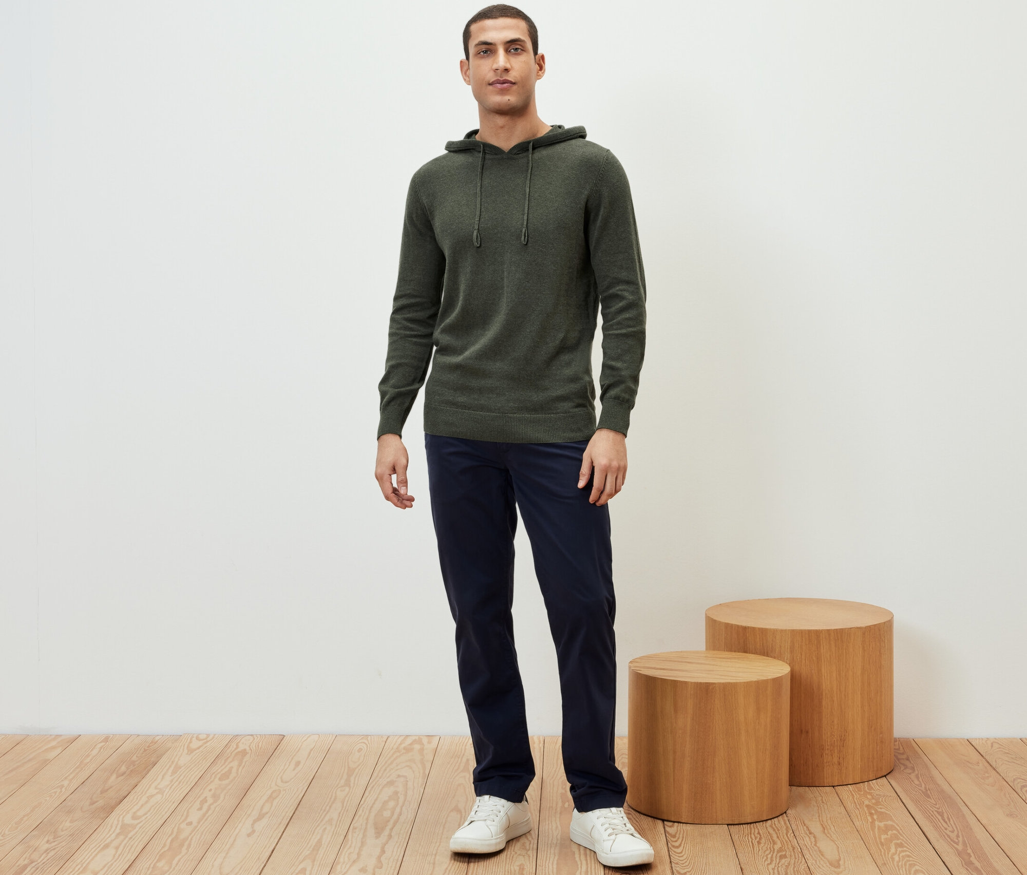 Un homme debout portant un sweatshirt à capuche en maille, un pantalon bleu marine et des baskets blanches.