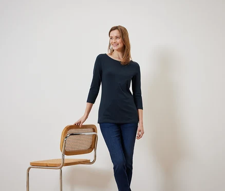 Une femme souriante portant un chemisier bleu marine à manches trois-quarts est appuyée contre une chaise en bois.