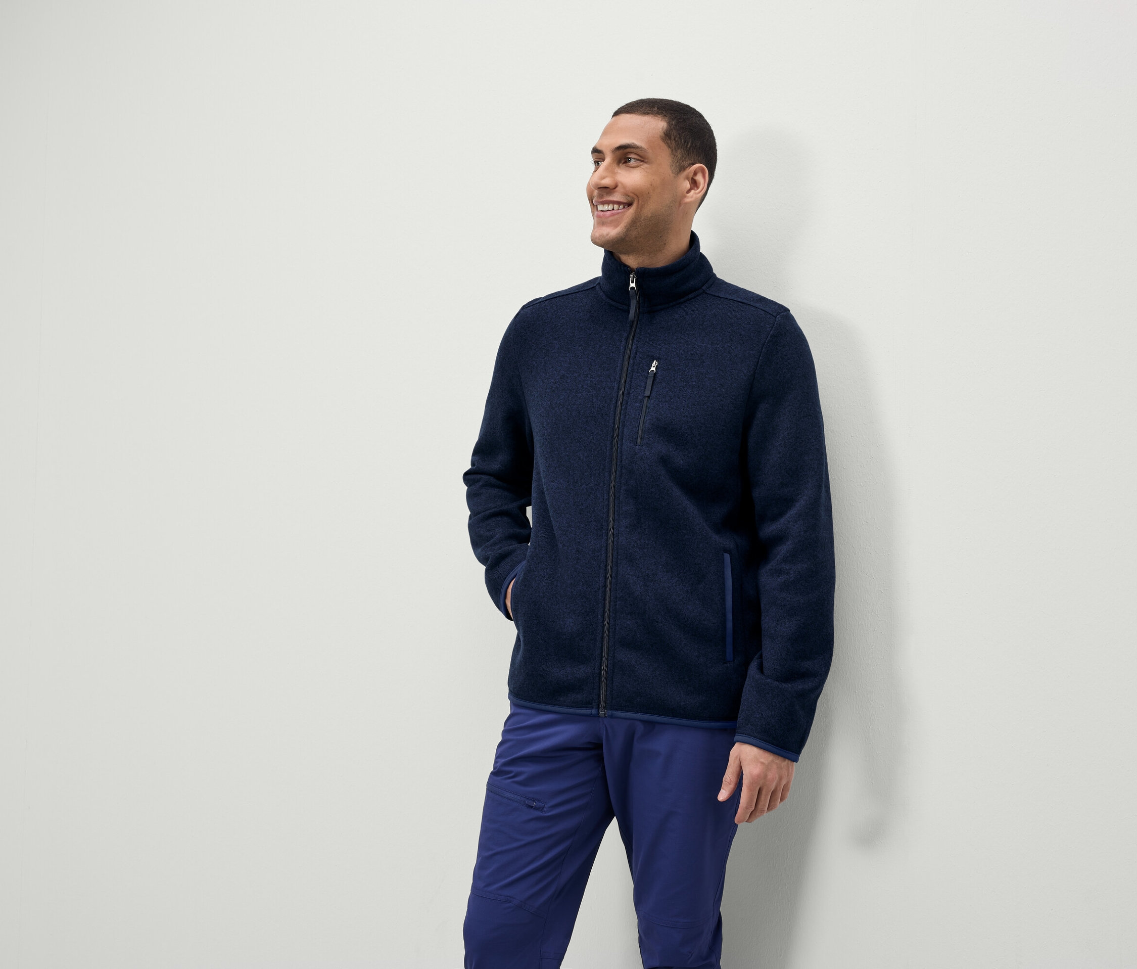 Un homme sourit et regarde sur le côté, il porte une veste polaire bleue zippée et un pantalon bleu.