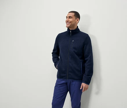 Un homme sourit et regarde sur le côté, il porte une veste polaire bleue zippée et un pantalon bleu.