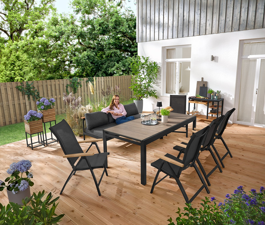 Une femme est assise sur un banc de jardin 2-en-1 ‒ une chaise longue «
Gefunden». À l’arrière-plan se trouvent 2 colonnes à plantes «
Tinus
». Sur la terrasse se trouvent également un chariot de grill avec plan de travail en polyciment et une table de jardin extensible avec plateau Duraboard.