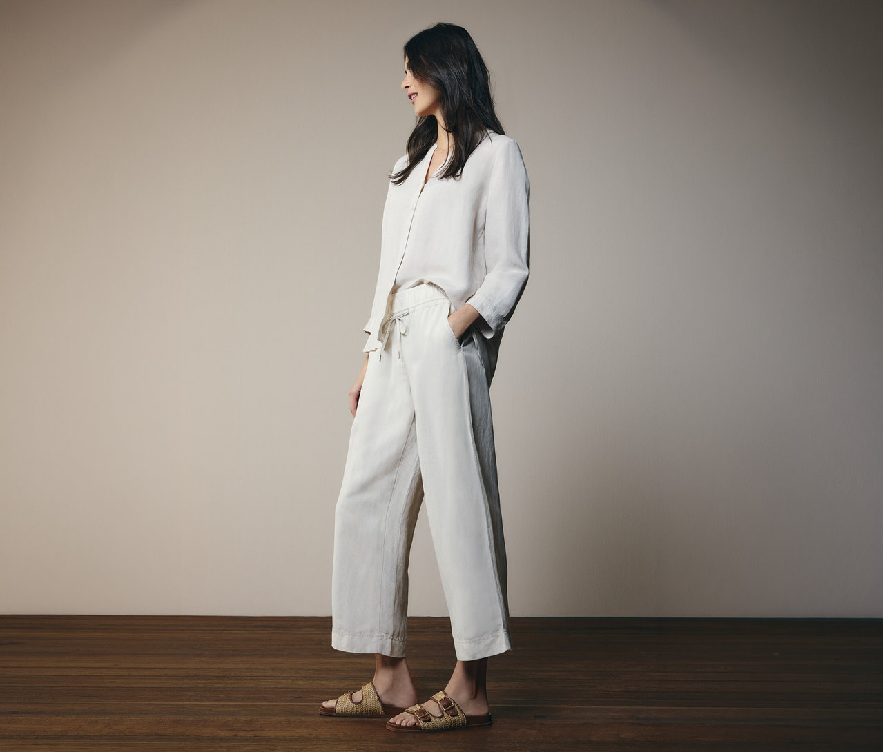 Femme en tenue de lin clair avec chemise et pantalon, debout pieds nus sur un plancher en bois.