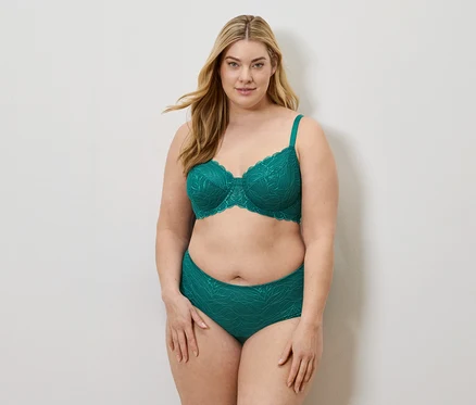 Une femme blonde porte un soutien-gorge émeraude et une culotte assortie à motif de feuilles sur fond blanc.