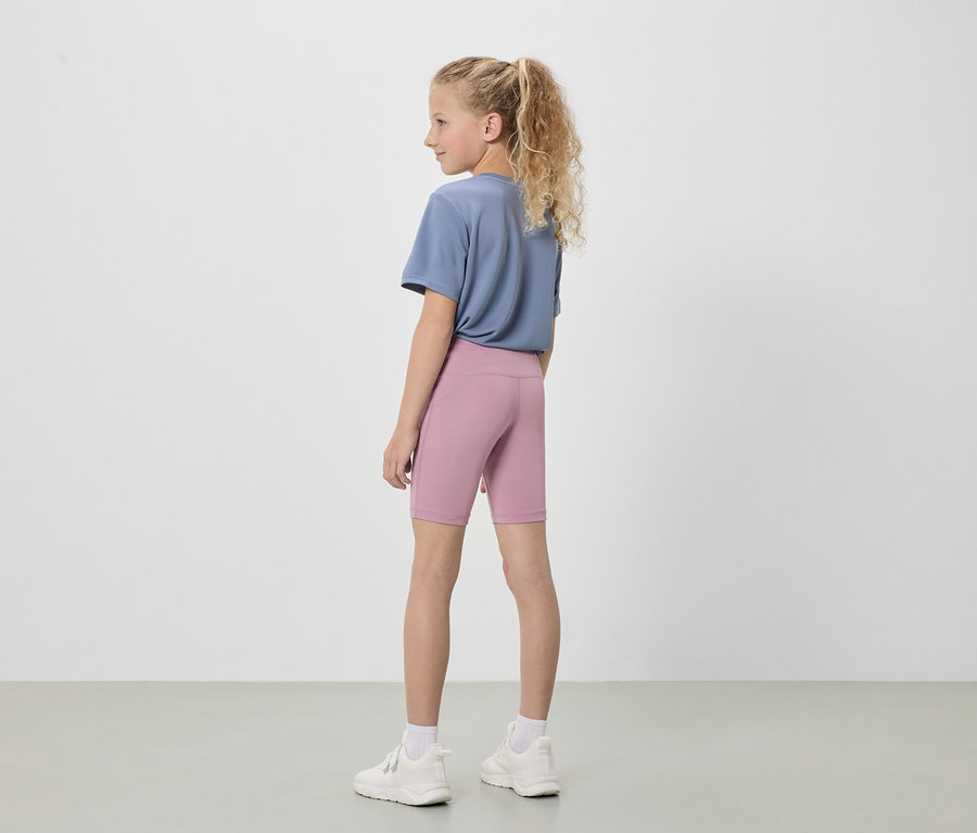 Une jeune fille blonde, vue de dos, porte un t-shirt bleu, un legging court rose et des baskets blanches sur un fond blanc.