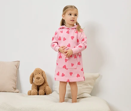 Une petite fille blonde avec des couettes se tient sur un lit et porte une robe à capuche rose avec des cœurs rouges. Un chien en peluche est assis à côté d'elle.