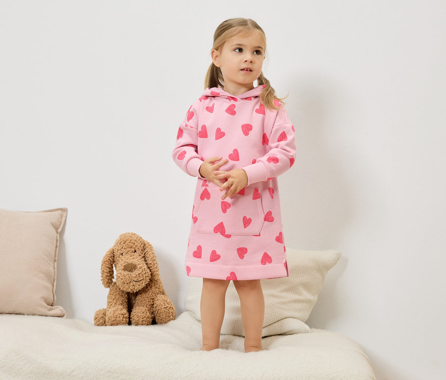 Une petite fille blonde avec des couettes se tient sur un lit et porte une robe à capuche rose avec des cœurs rouges. Un chien en peluche est assis à côté d'elle.