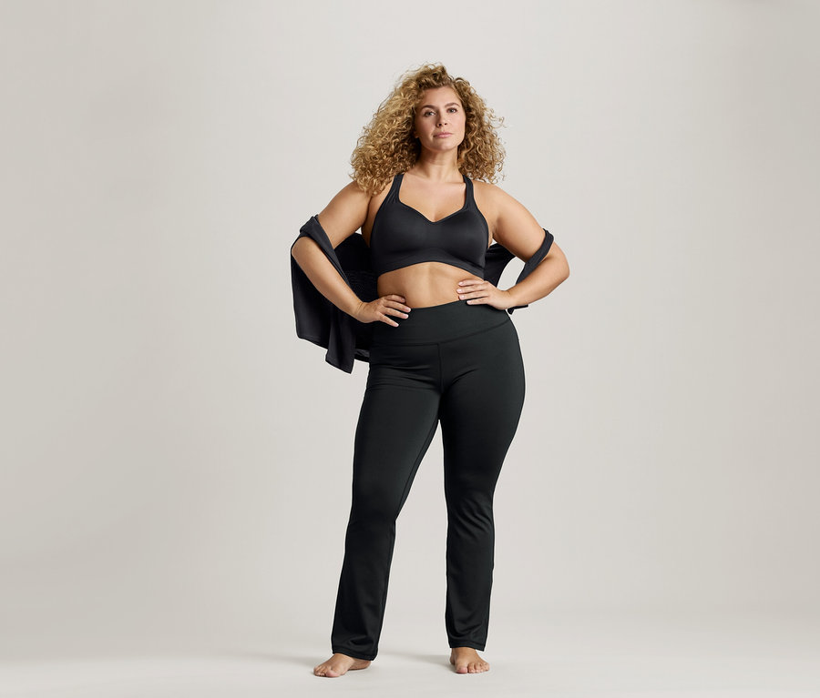 Femme aux cheveux bouclés portant un soutien-gorge de sport noir et un legging, avec une veste sur les épaules et les mains sur les hanches.