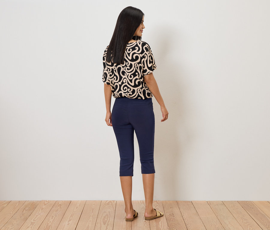 Femme vue de dos portant un haut à motifs, un pantacourt bleu et des sandales devant un mur blanc.