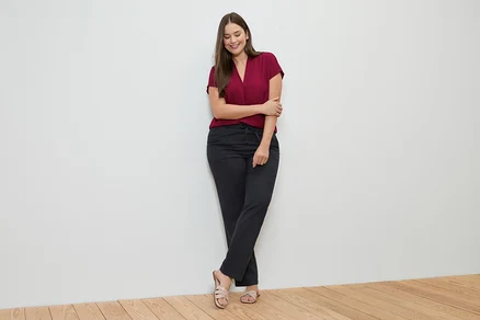 Femme appuyée contre un mur, portant un haut bordeaux, un pantalon en lin noir et des sandales.