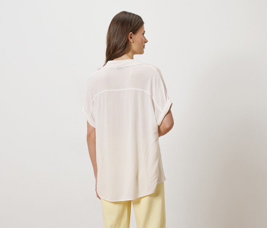 Une femme aux cheveux bruns porte une chemise claire et un pantalon jaune, vue de dos.