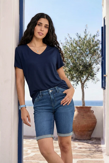 Une femme s'appuie contre un mur, porte un t-shirt bleu marine, un bermuda en jean, des bagues pavées bleues et transparentes et une montre bracelet avec un bracelet en cuir.
