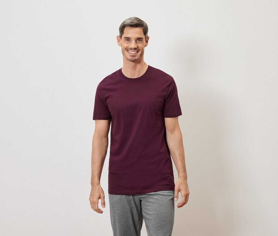 Un homme aux cheveux grisonnants sourit, vêtu d'un t-shirt bordeaux et d'un pantalon gris sur fond blanc.