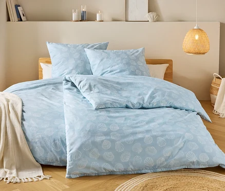 Une chambre avec une literie bleu clair à motifs de coquillages, un tabouret en bois avec une couverture et une lampe tressée.