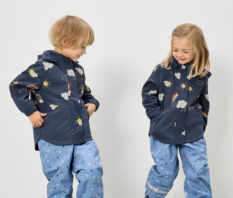 Deux enfants portant des vestes bleues à motifs arc-en-ciel, nuages et soleil, et des pantalons bleus à pois se tiennent devant un fond blanc.