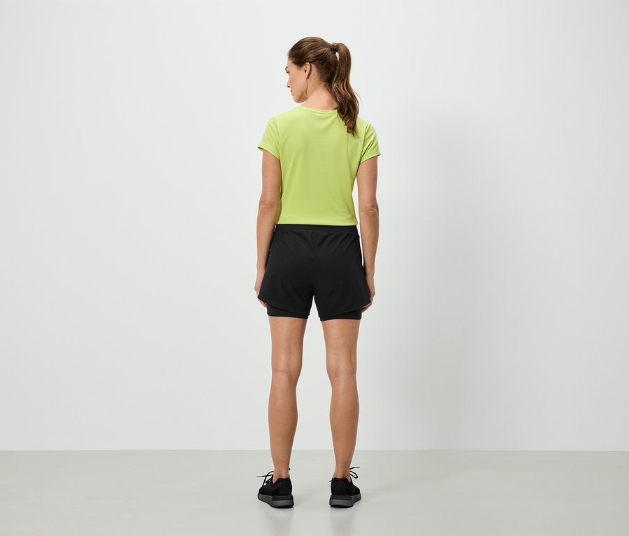 Une femme se tient dos à la caméra, portant un t-shirt vert clair et un short noir.