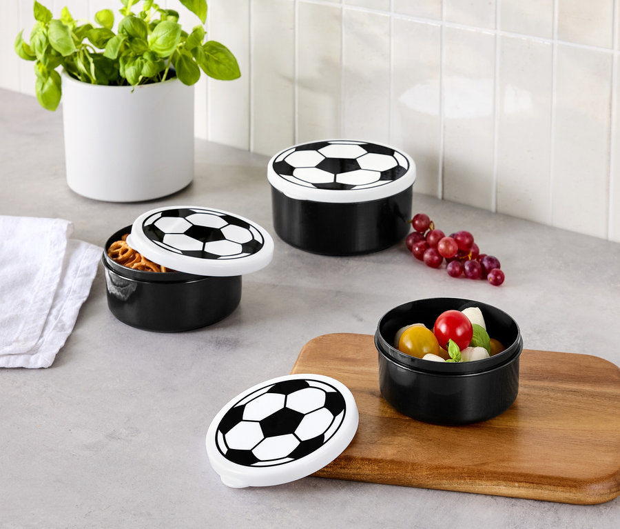 Trois boîtes de conservation noires avec des couvercles de football sont posées sur un plan de travail de cuisine, une boîte est ouverte et remplie de bretzels, une avec des tomates et de la mozzarella.