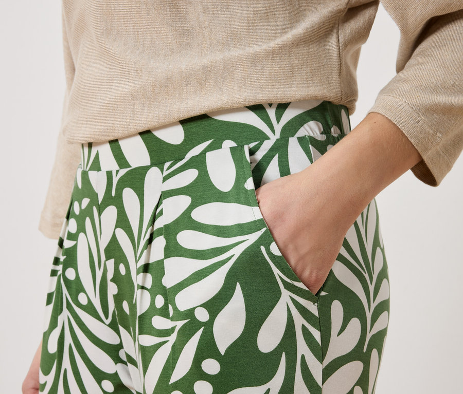 Une personne porte un chemisier beige et un pantalon vert avec un motif de feuilles blanches. Une main est dans la poche du pantalon.