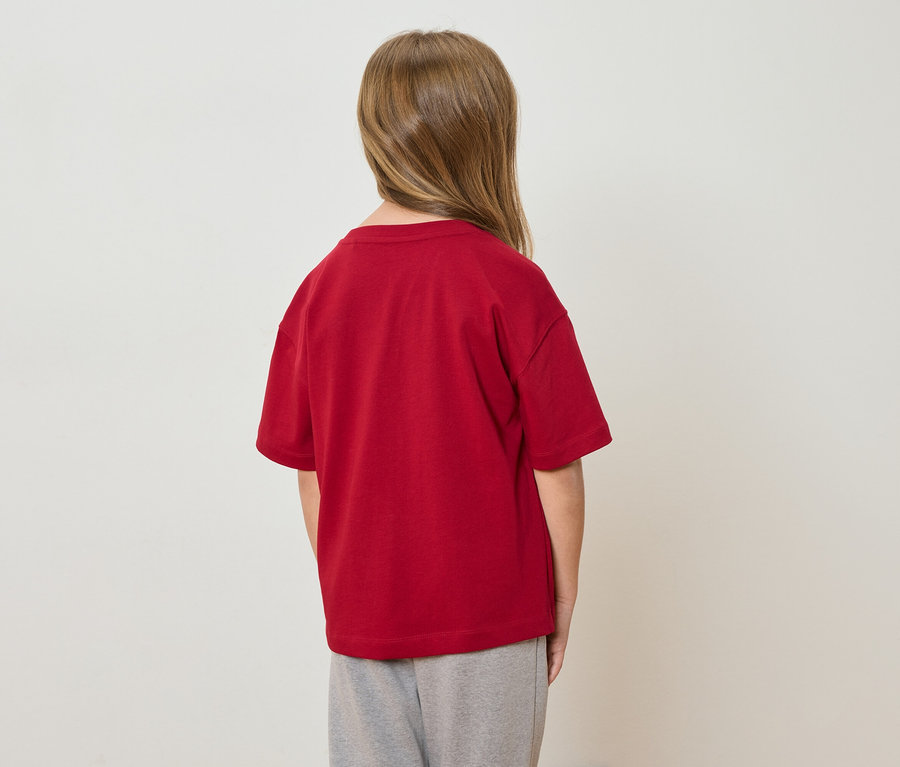 Un enfant aux cheveux blonds se tient dos à la caméra, portant un t-shirt rouge et un pantalon gris.