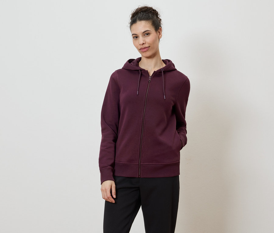 Une femme portant un chignon porte un sweat à capuche bordeaux zippé et un pantalon noir sur fond blanc.