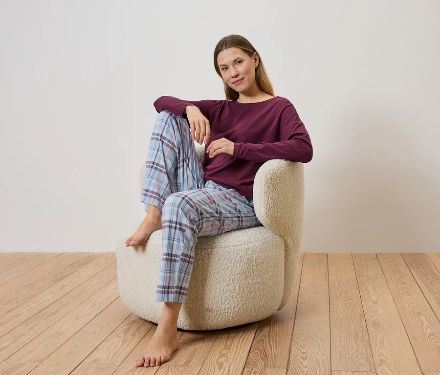Une femme est assise pieds nus dans un pantalon de pyjama à carreaux et un haut bordeaux dans un fauteuil beige sur un plancher en bois.
