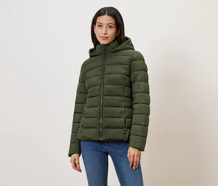 Une femme porte une doudoune vert olive à capuche et fermeture éclair, avec un jean bleu sur fond clair.