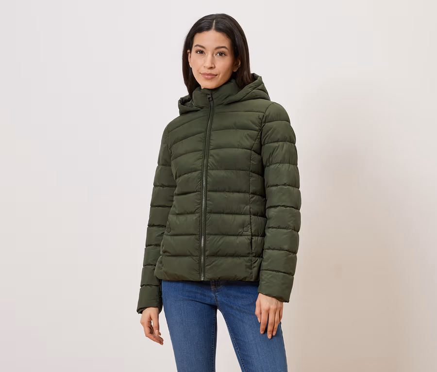 Une femme porte une doudoune vert olive à capuche et fermeture éclair, avec un jean bleu sur fond clair.
