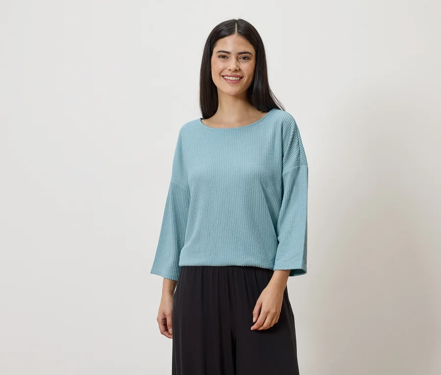 Une femme aux longs cheveux foncés porte un haut bleu clair à manches longues et un pantalon noir sur un fond blanc.