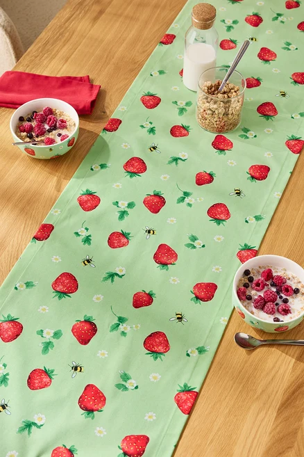 Sur une table en bois se trouve un chemin de table à motifs de fraises, dessus des bols de céréales, un verre de muesli et une bouteille de lait.
