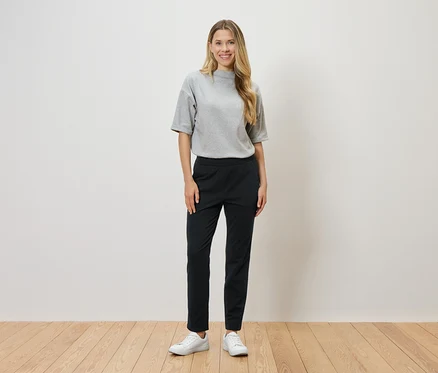 Une femme aux cheveux blonds se tient devant un mur blanc et porte un t-shirt gris, un pantalon noir et des baskets blanches.