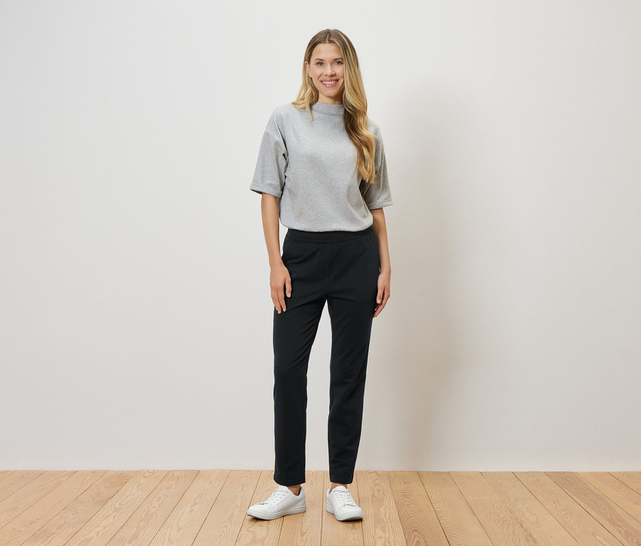 Une femme aux cheveux blonds se tient devant un mur blanc et porte un t-shirt gris, un pantalon noir et des baskets blanches.