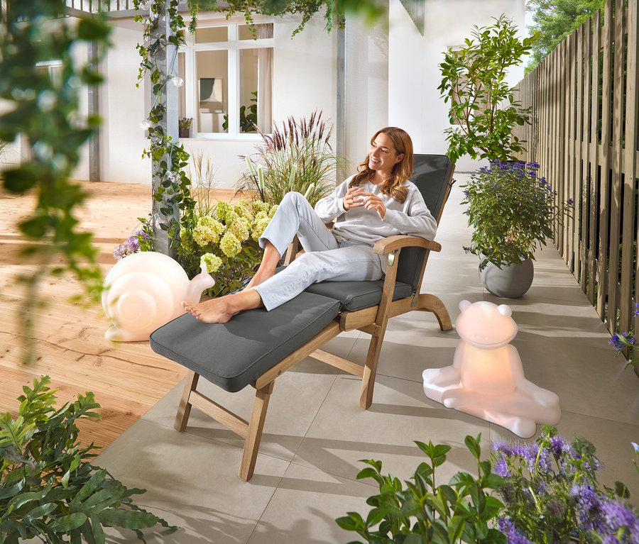 Une femme est assise sur une chaise de pont avec repose-pieds extensible. Dans le jardin, on voit une lampe solaire Premium « Escargot », une lampe solaire Premium « Grenouille » et une guirlande lumineuse solaire « Ampoules ».