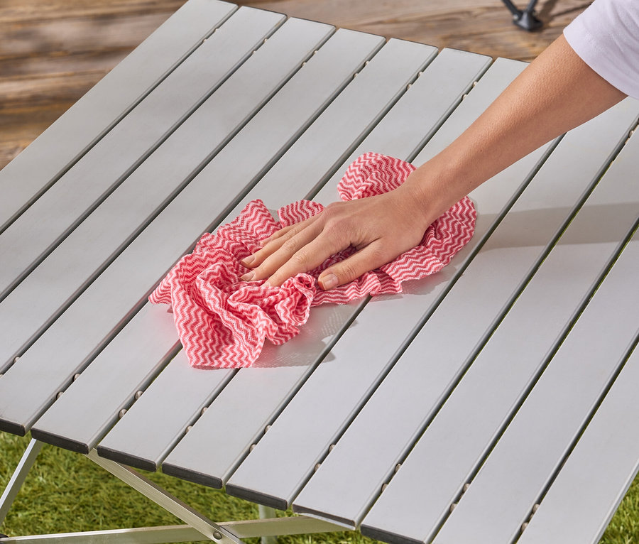 Une main essuie une table de camping grise avec un chiffon rouge et blanc à carreaux.