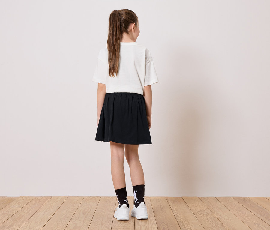 Vue de dos d'une jeune fille avec une queue de cheval, un t-shirt blanc, une jupe noire et des chaussettes avec une figurine blanche debout sur un plancher en bois.