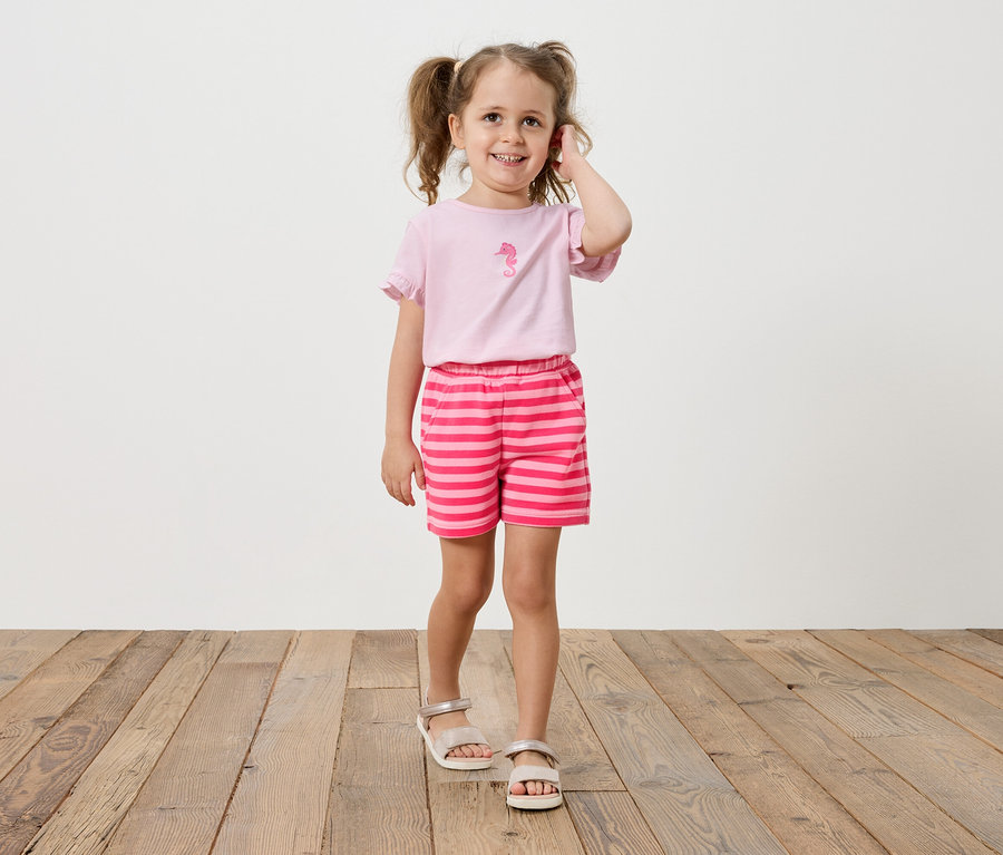 Fillette avec des tresses porte un t-shirt rose avec un imprimé hippocampe et un short rayé.