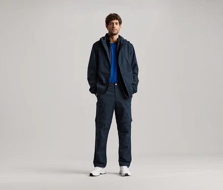 Un homme se tient debout, les mains dans les poches d'une veste bleu marine et d'un pantalon assorti, portant un t-shirt bleu et des baskets blanches.