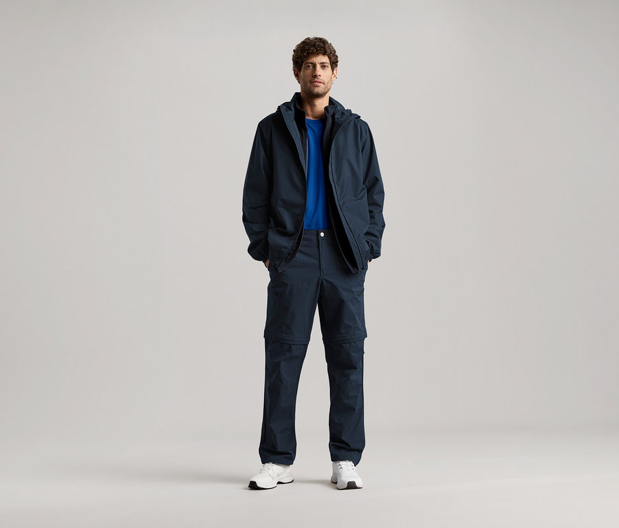 Un homme se tient debout, les mains dans les poches d'une veste bleu marine et d'un pantalon assorti, portant un t-shirt bleu et des baskets blanches.