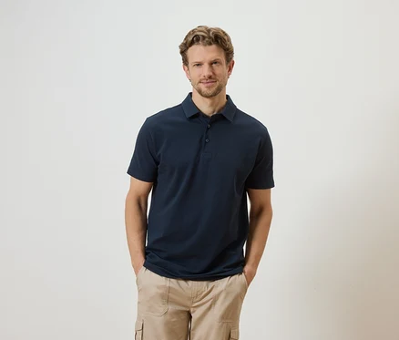Un homme aux cheveux blonds et à la barbe, vêtu d'un polo bleu marine et d'un pantalon cargo beige, pose devant un fond blanc.