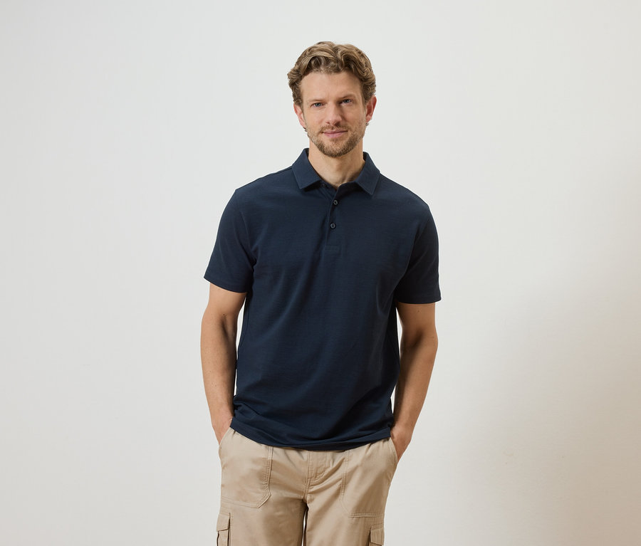 Un homme aux cheveux blonds et à la barbe, vêtu d'un polo bleu marine et d'un pantalon cargo beige, pose devant un fond blanc.