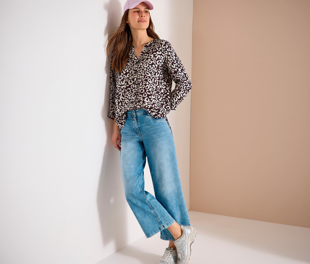 Une femme avec une casquette rose, un chemisier à motifs, un jean bleu et des baskets argentées est appuyée contre un mur.